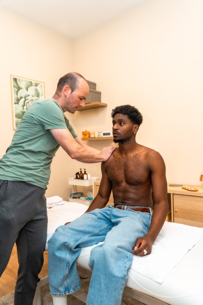 Physical therapist massaging a young black man's shoulder while he sits on an examination table, focusing on rehabilitation and muscle relief in a wellness setting
